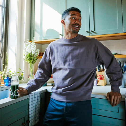Man standing in a kitchen holding a glass of juice, wearing a gray sweater and blue shorts.
